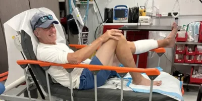 John Schweizer sentado numa cama de hospital após a mordida de tubarão nas Bahamas, com a parte inferior da perna esquerda enfaixada