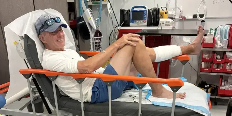 John Schweizer sentado numa cama de hospital após a mordida de tubarão nas Bahamas, com a parte inferior da perna esquerda enfaixada