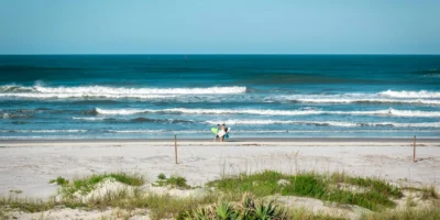 Duas pessoas com pranchas de surfe em New Smyrna Beach, com ondas ao fundo